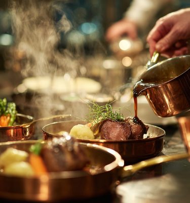 Patrick Coudert gießt aromatische Sauce aus einem Kupfertopf über ein Stück Fleisch auf einem edel angerichteten Teller mit Gemüse, umgeben von dampfenden Gerichten in Kupferpfannen.