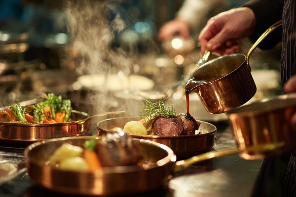 Patrick Coudert gießt aromatische Sauce aus einem Kupfertopf über ein Stück Fleisch auf einem edel angerichteten Teller mit Gemüse, umgeben von dampfenden Gerichten in Kupferpfannen.
